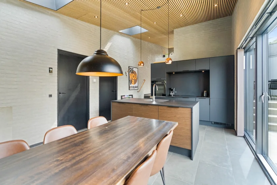 Un comedor moderno se abre a una cocina, con una mesa larga de madera junto a grandes ventanas.