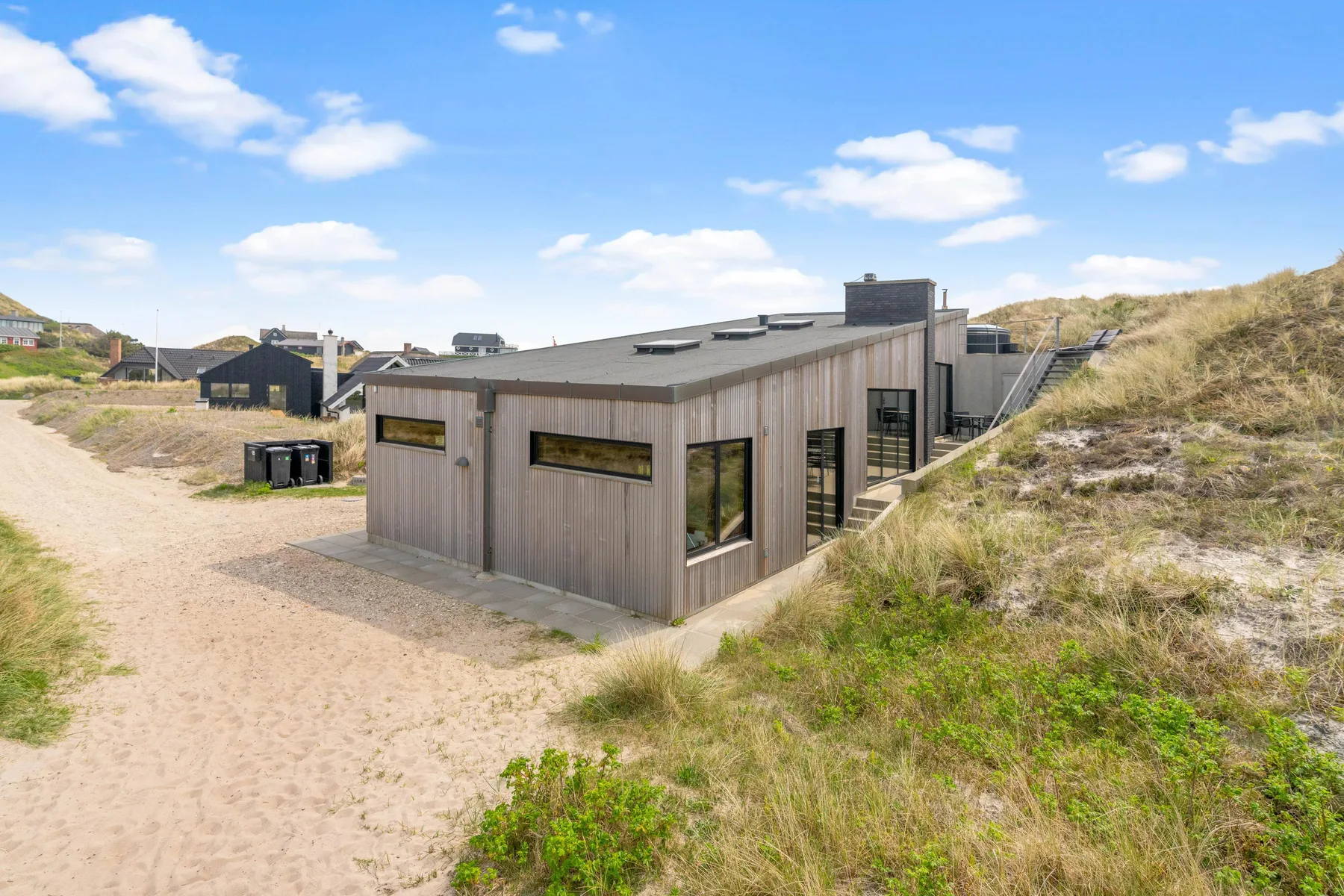 Una casa moderna está junto a caminos de arena y dunas cubiertas de hierba bajo un cielo azul.