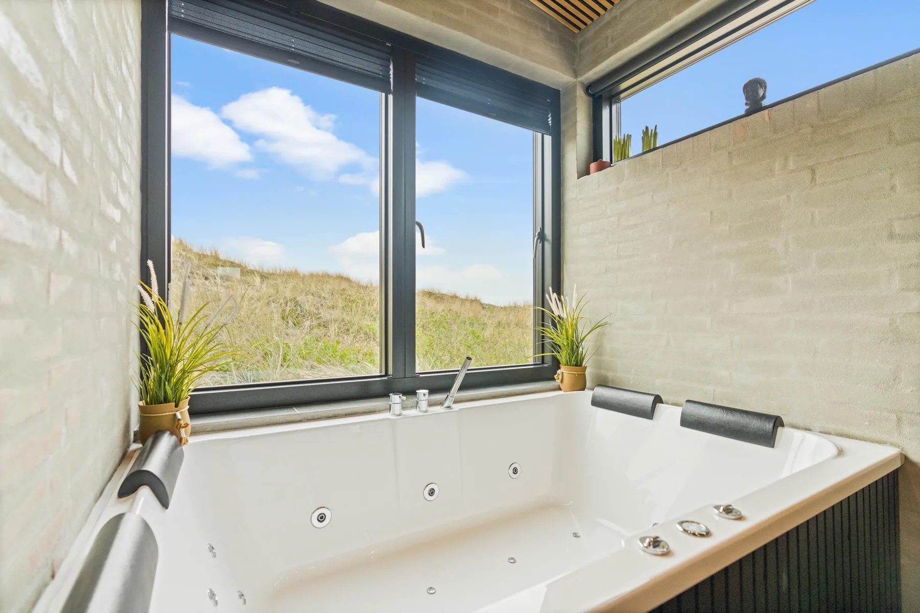 Una bañera de hidromasaje empotrada está bajo grandes ventanas con vistas a dunas cubiertas de hierba.