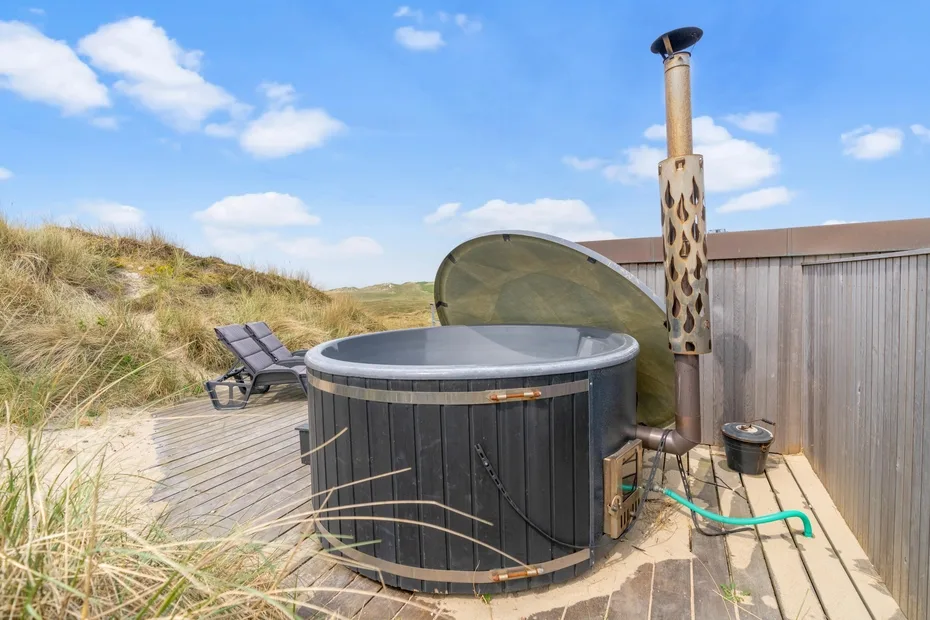 Una bañera de hidromasaje exterior está sobre una terraza de madera junto a dunas cubiertas de hierba.