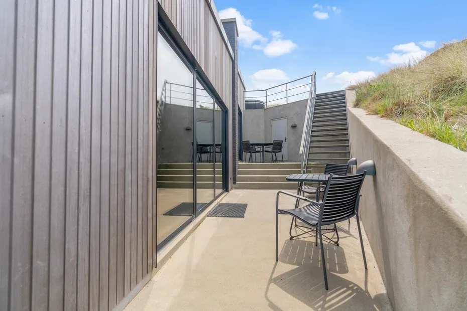 Una terraza exterior soleada tiene asientos junto a un edificio moderno y unas escaleras.