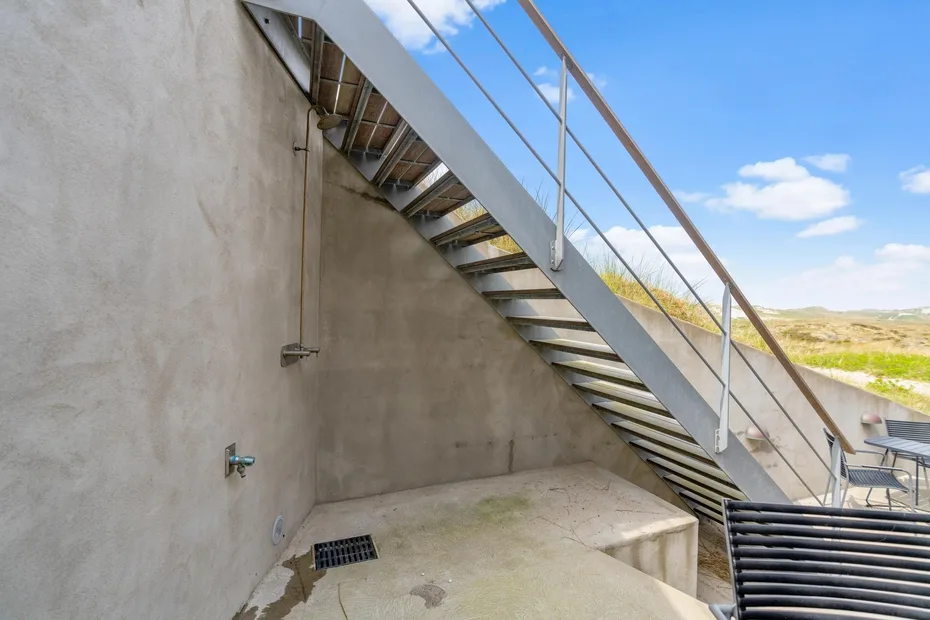 Una escalera exterior desciende junto a un patio de hormigón, con campo abierto al fondo.