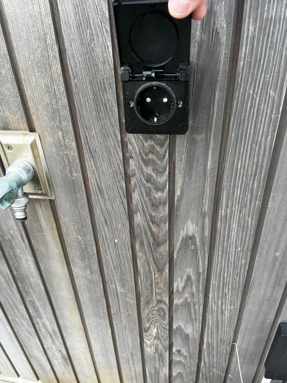 Primer plano de un tomacorriente con la tapa protectora abierta junto a un grifo metálico.