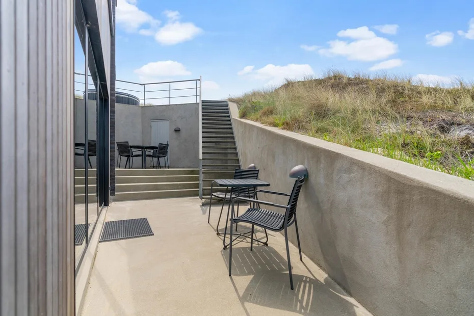 Una terraza exterior pavimentada tiene asientos metálicos junto a una pendiente con hierba y escaleras.
