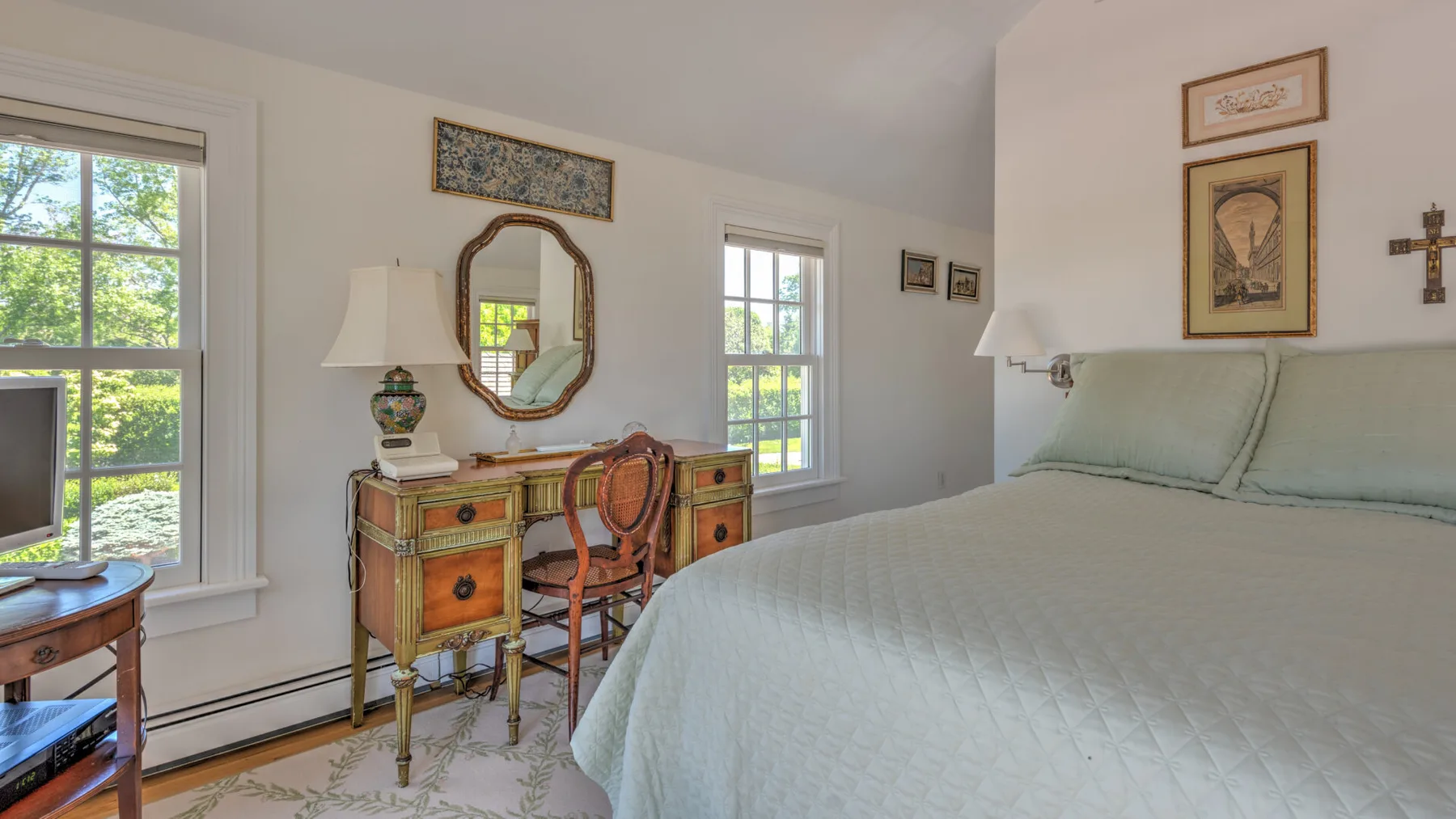 A bright bedroom with a bed, antique dresser, and windows overlooking greenery.