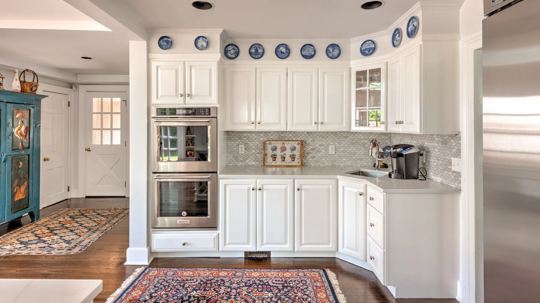 A bright kitchen with white cabinets, built-in ovens, and a tiled backsplash.