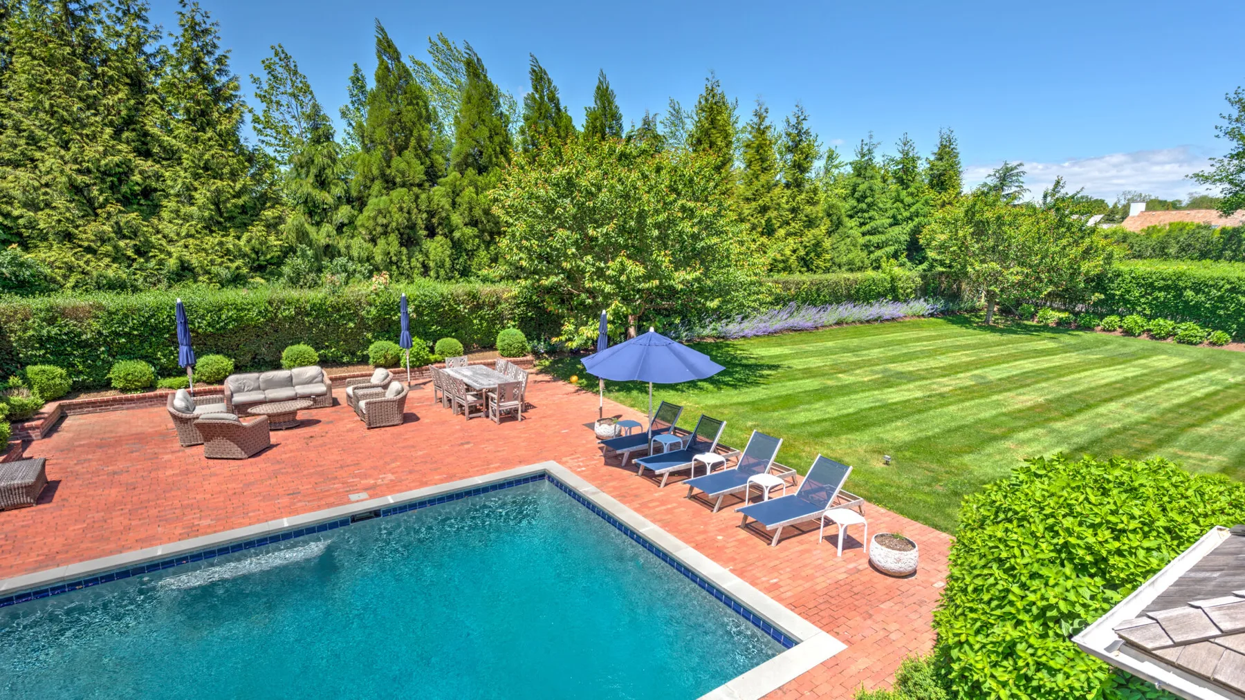 A backyard pool borders a brick patio with outdoor seating and a striped lawn.