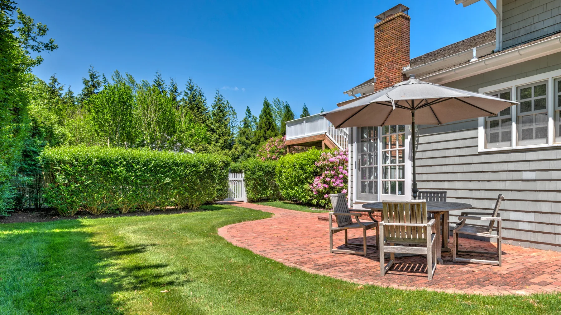 A brick patio beside a house opens onto a green lawn and leafy hedges.
