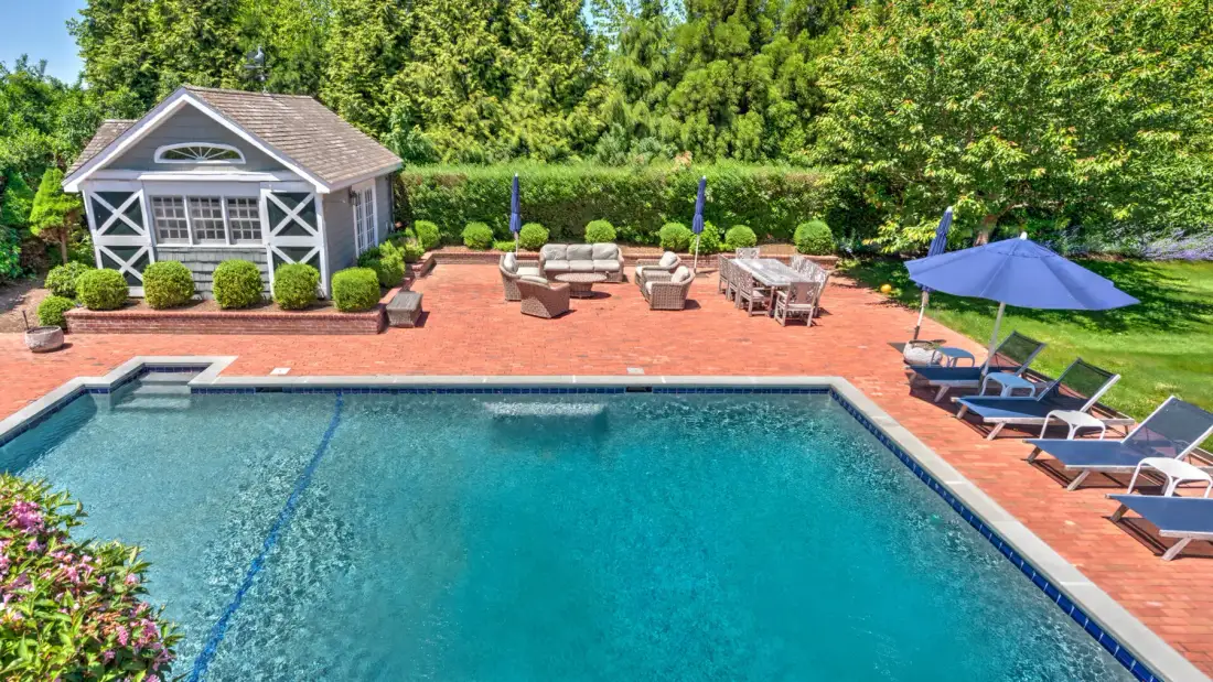 An outdoor swimming pool borders a brick patio with seating and umbrellas.