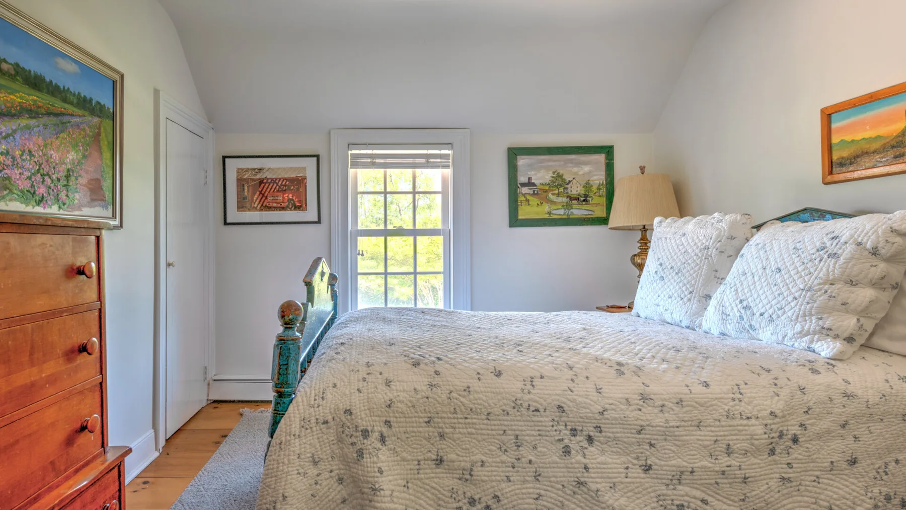 A bedroom with a bed covered in a light patterned quilt and framed artwork.
