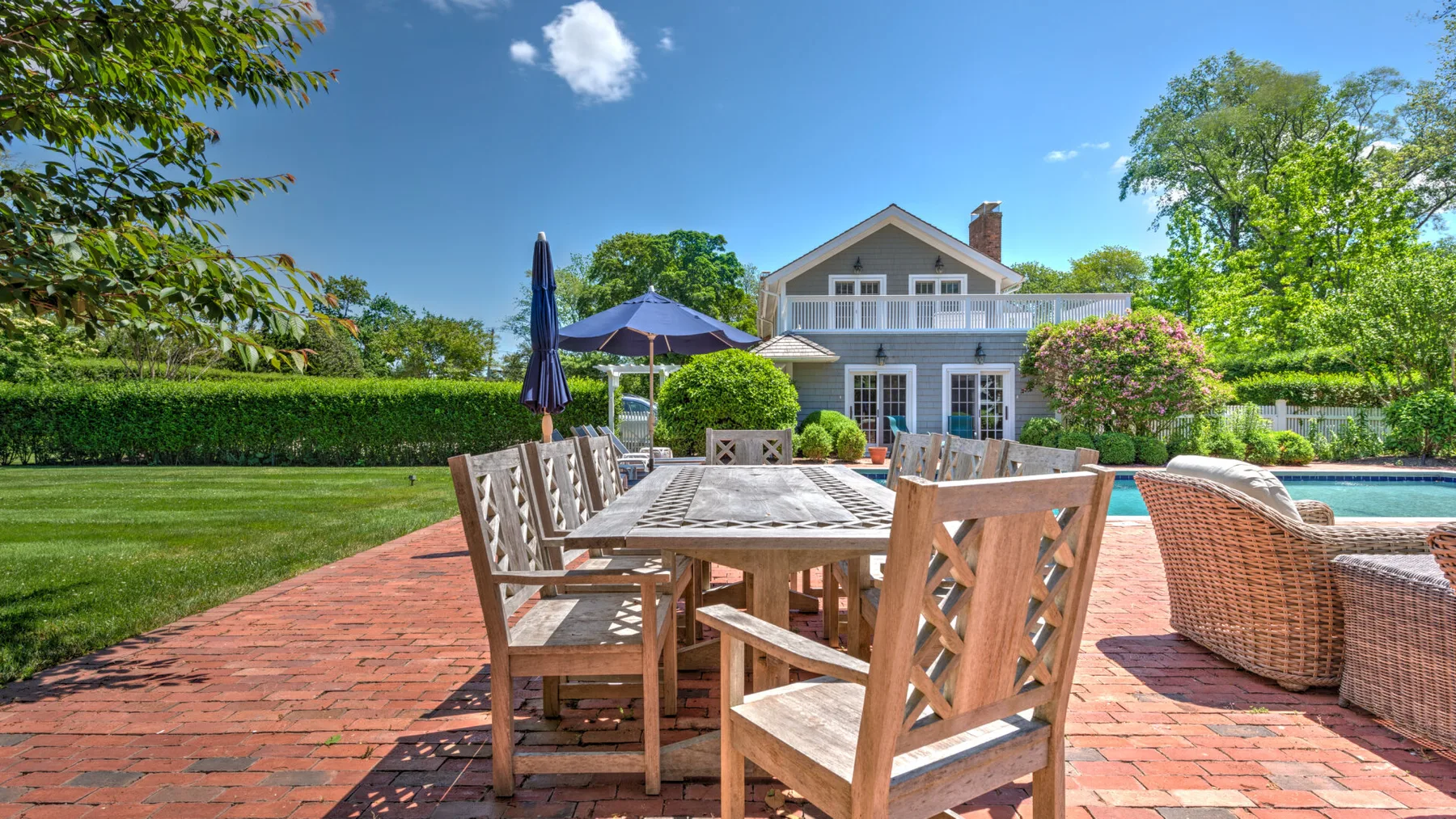 Una zona de comedor al aire libre con una mesa de madera junto a una casa y una piscina.