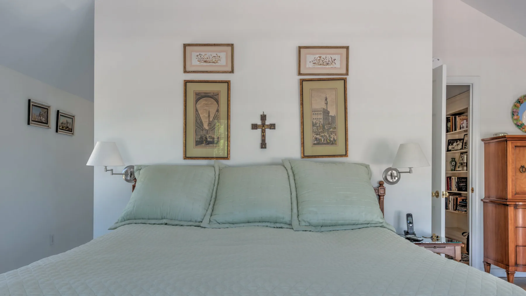 A bedroom with a neatly made bed beneath framed pictures and wall lamps.
