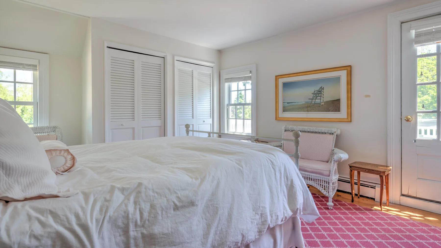 A bright bedroom with a large bed, pale walls, and a pink patterned rug.