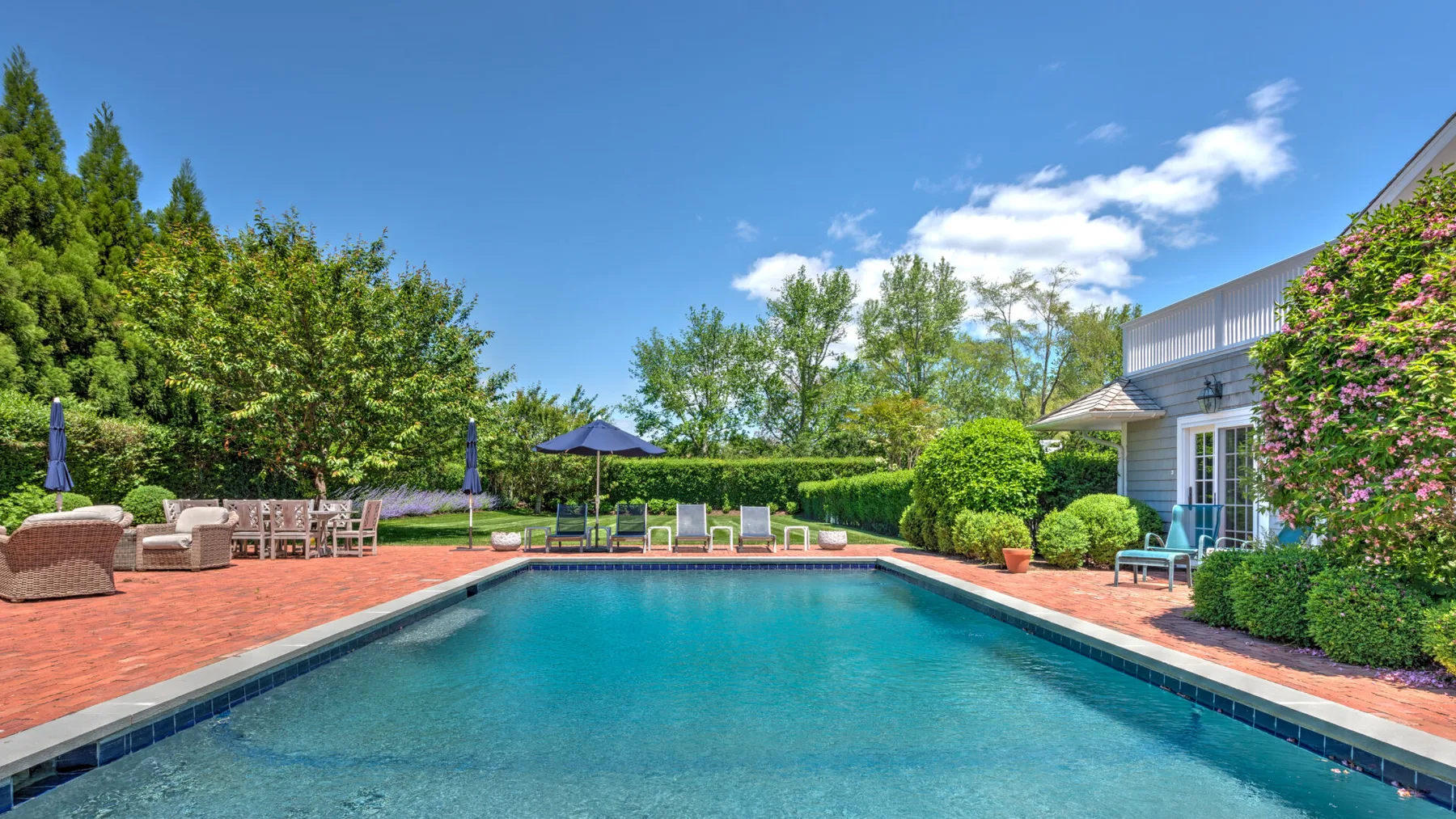 Piscina al aire libre junto a un patio pavimentado, con asientos y vegetación alrededor.