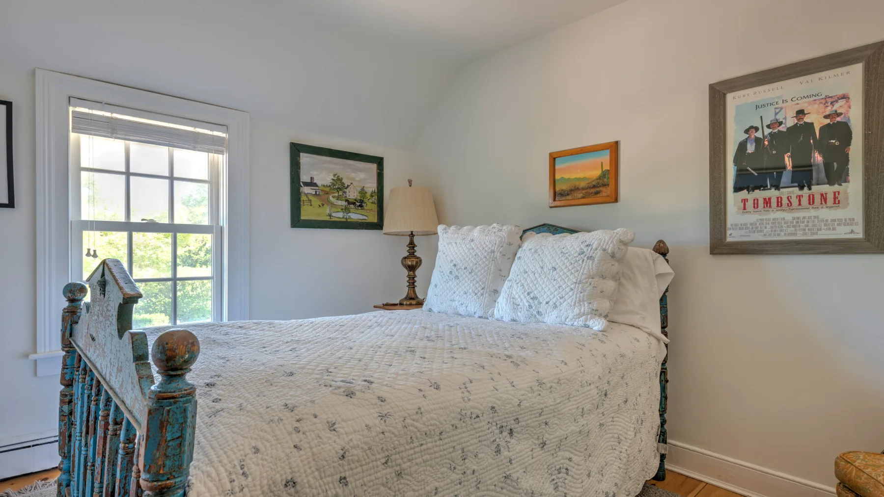 A bedroom with a bed beneath a window and framed artwork on the walls.