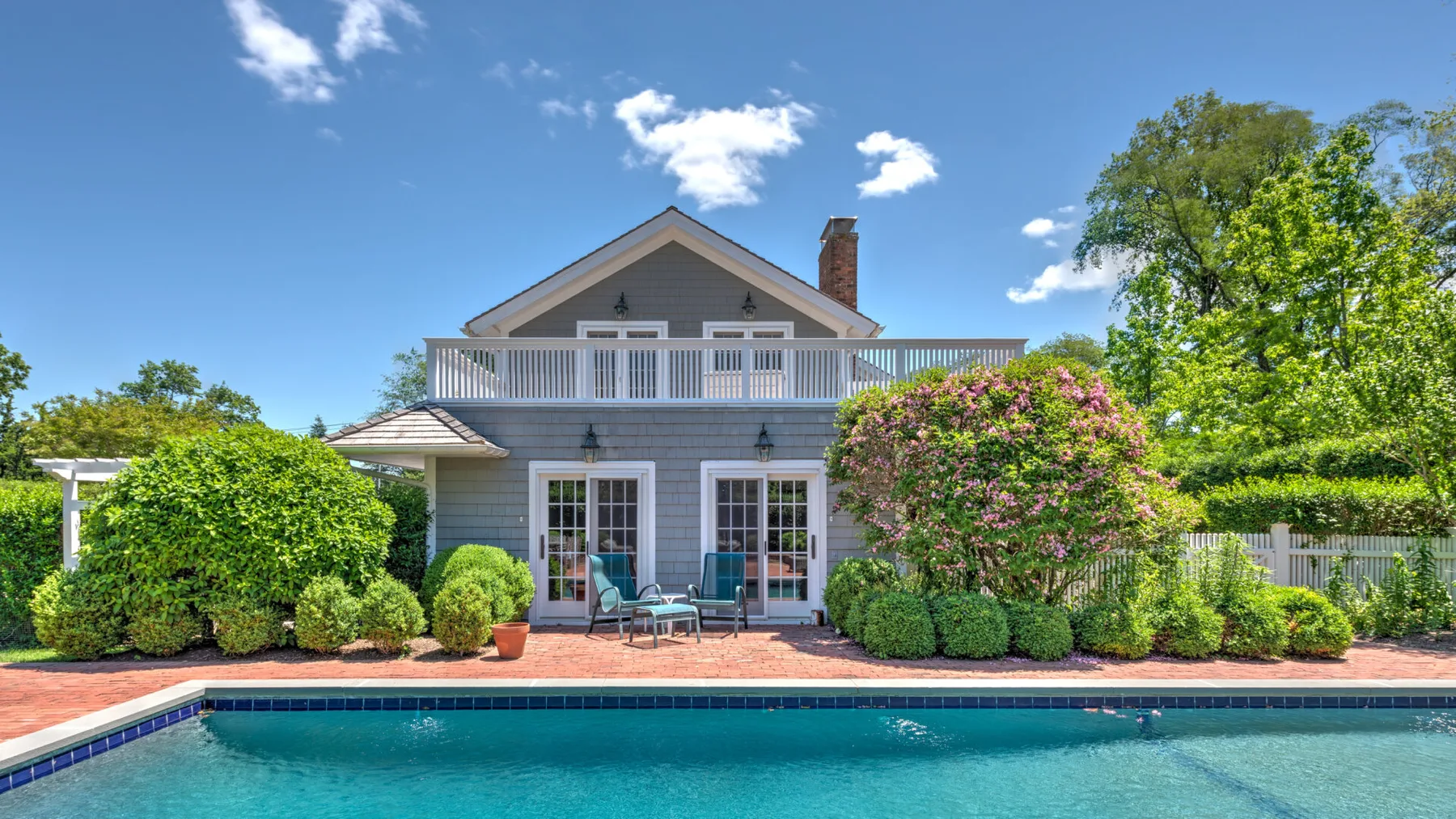 A landscaped house opens onto a swimming pool, with patio seating and flowering shrubs.