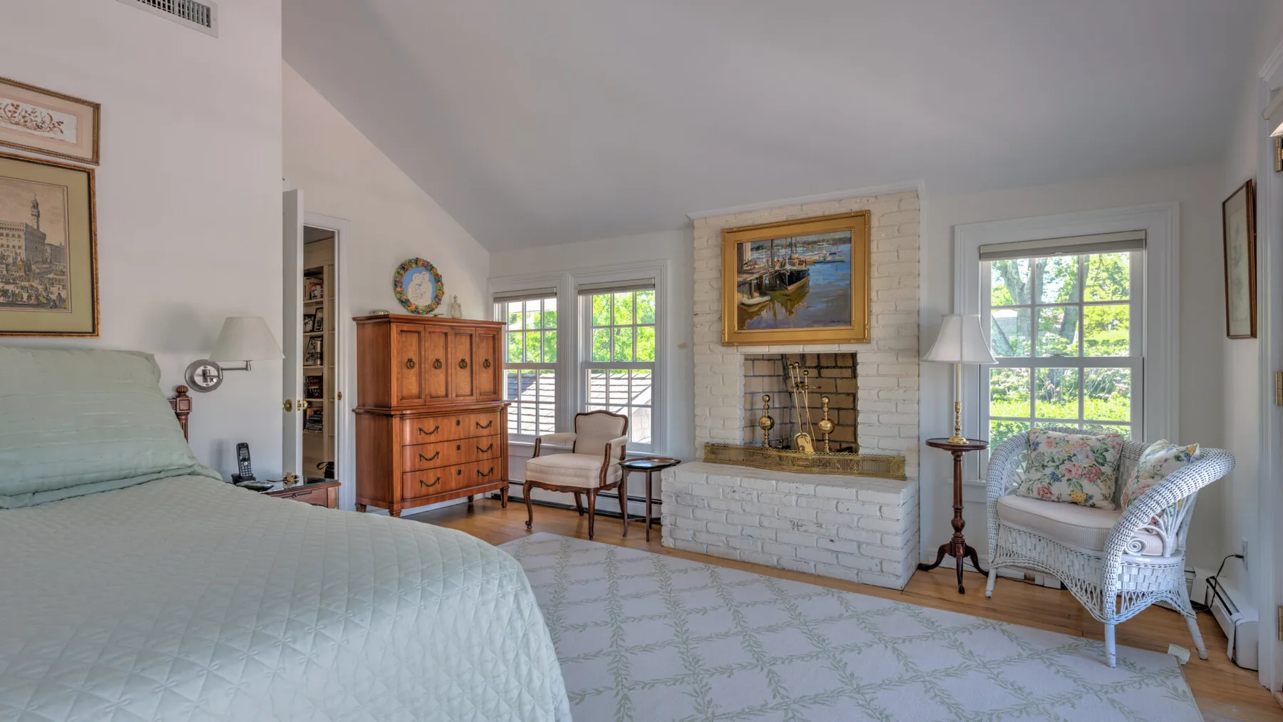 A bright bedroom with a bed, a brick fireplace, and seating by the windows.