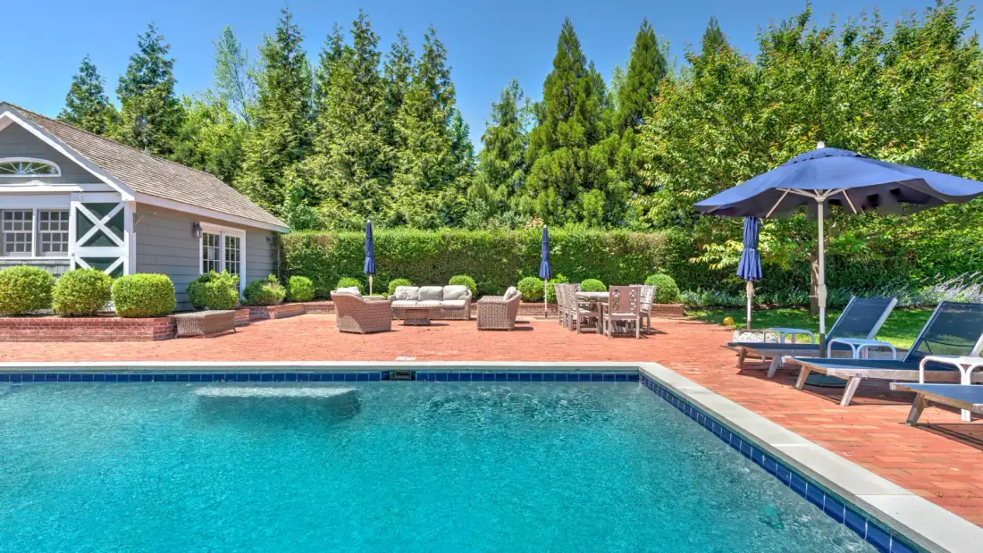 An outdoor swimming pool beside a paved patio with seating and umbrellas.
