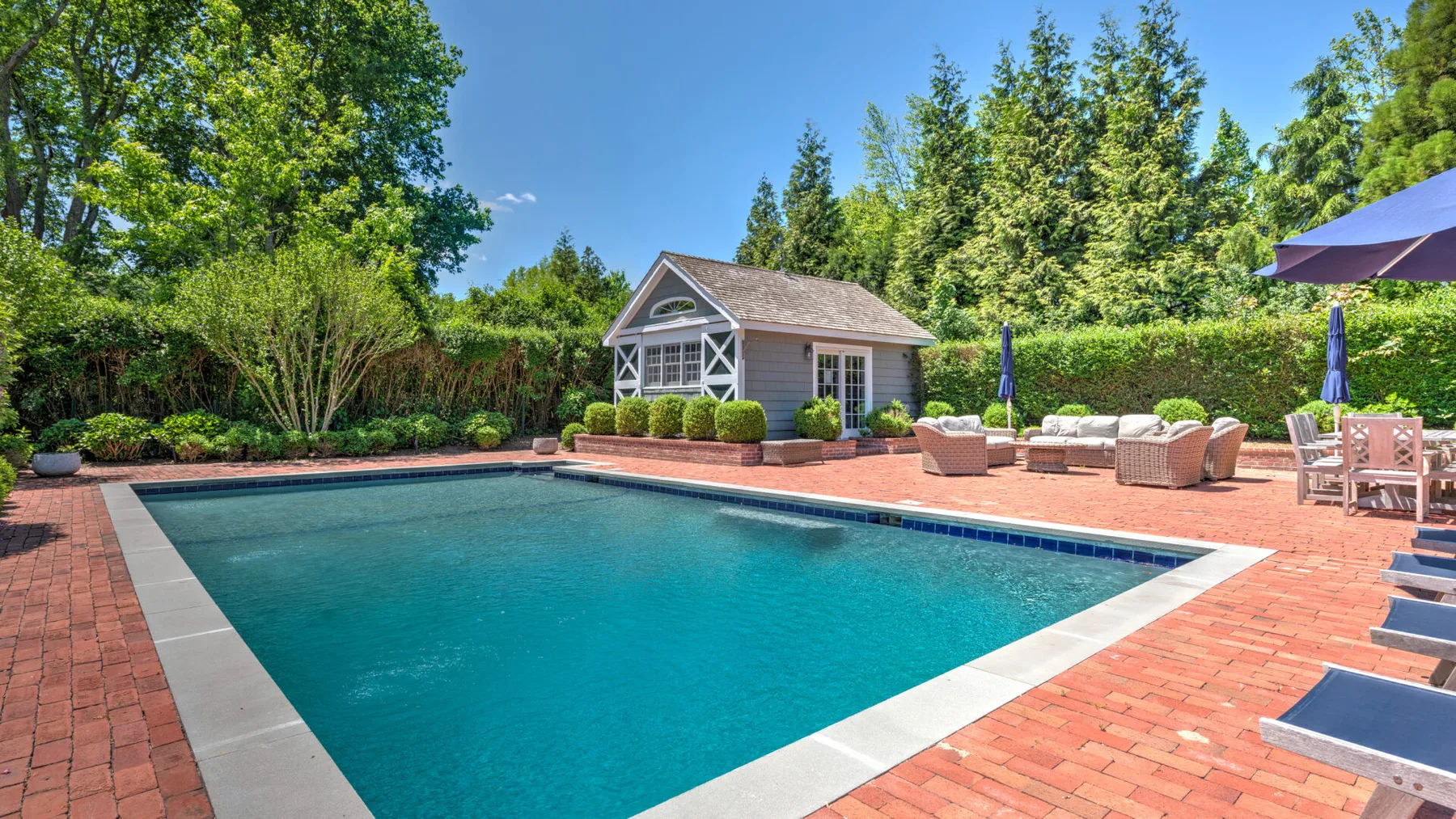 An outdoor swimming pool sits beside a brick patio and small garden building.