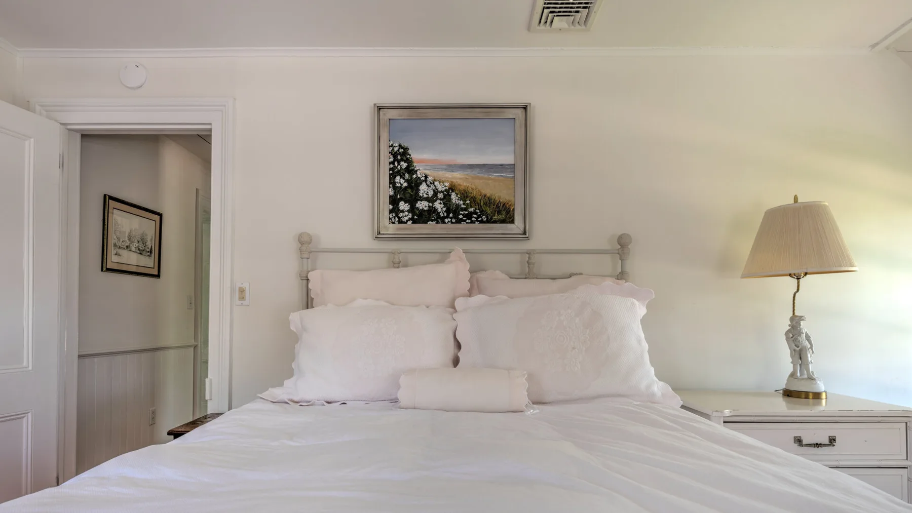 A bedroom with a neatly made bed and framed landscape artwork above it.