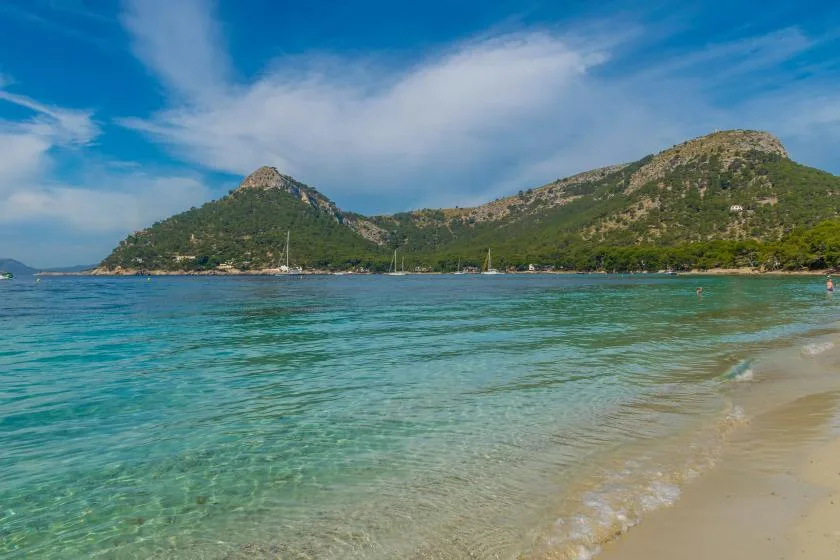 A sandy beach meets clear turquoise water, with green hills across the bay.