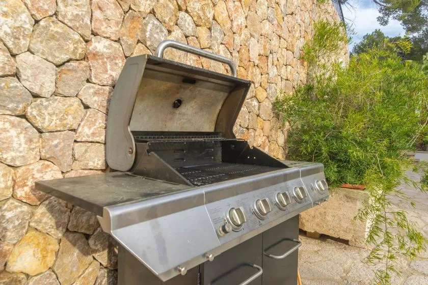 An outdoor gas grill stands beside a stone wall and greenery.