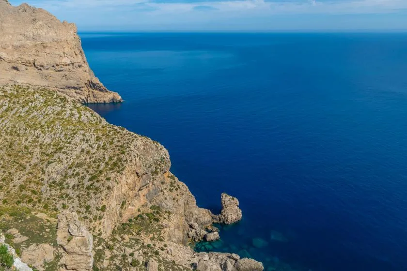 A rocky coastal cliff overlooks clear blue sea beneath a bright sky.
