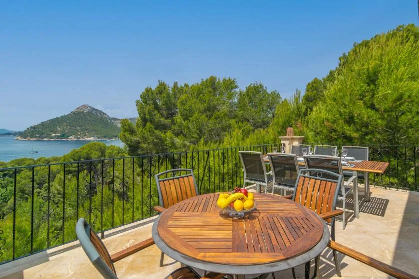 An outdoor terrace has a round wooden table and seating overlooking trees and the sea.