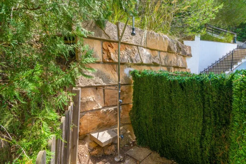 An outdoor shower stands beside a stone retaining wall and dense greenery.