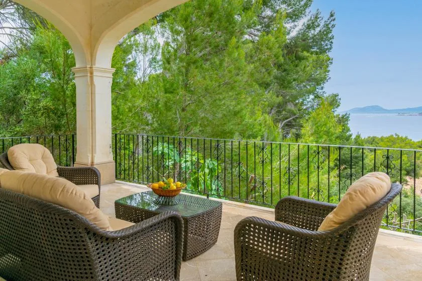 A covered terrace has wicker seating and a view of the water beyond the railing.
