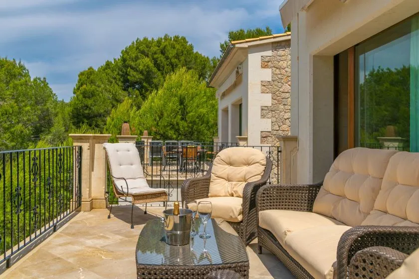 An outdoor terrace has cushioned seating and a glass-topped table overlooking green trees.