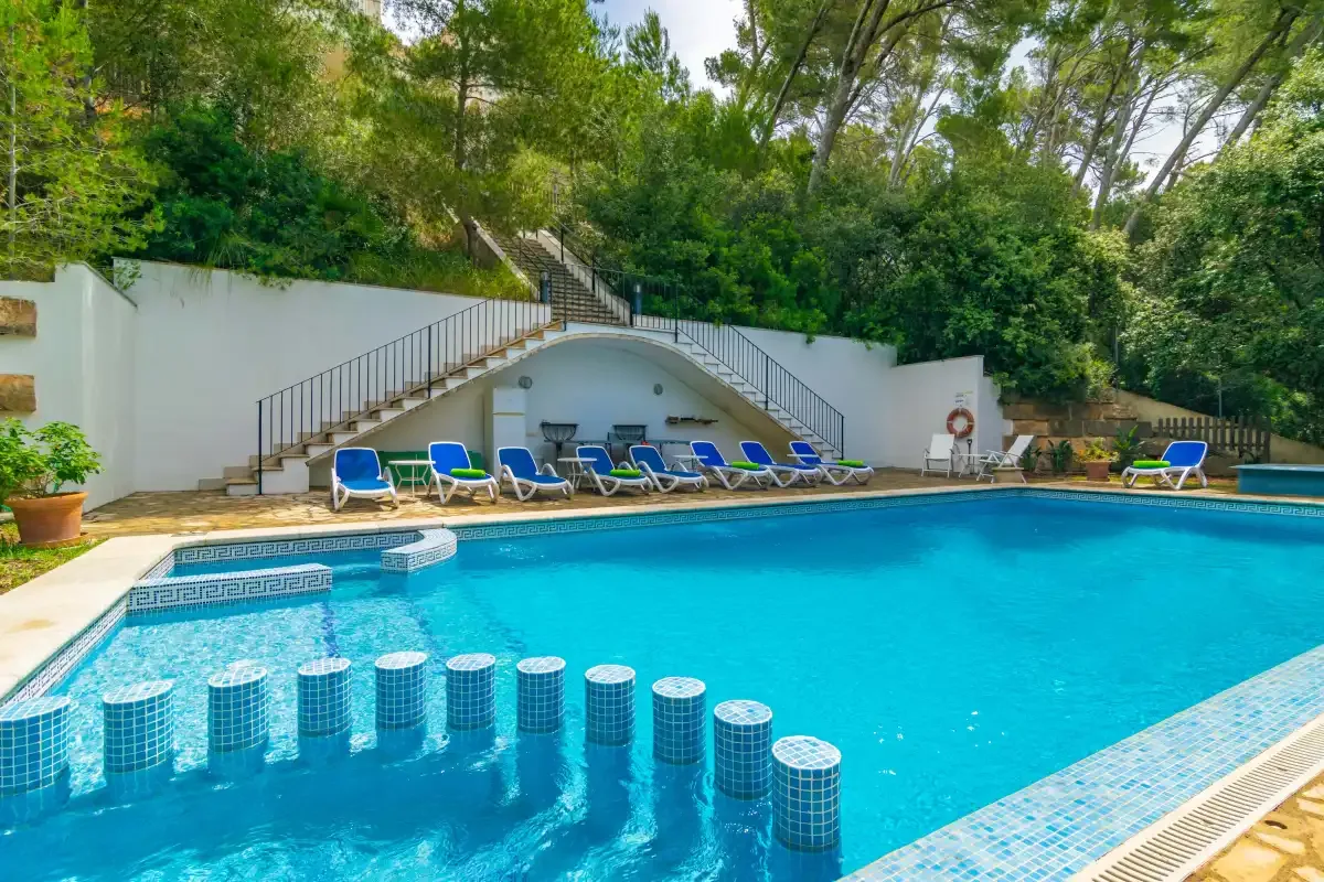 An outdoor swimming pool with blue loungers beside a white wall and trees.