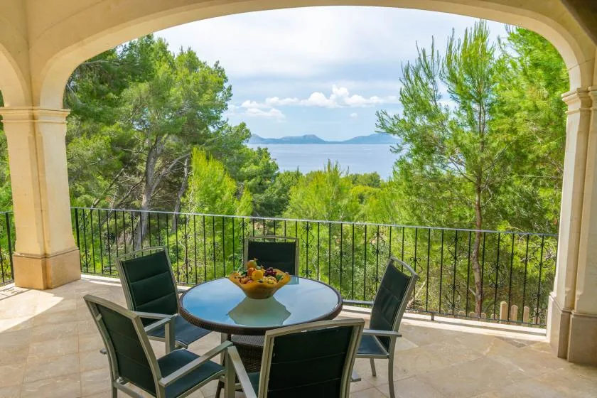 A covered terrace with a table and seating overlooks trees and the water.