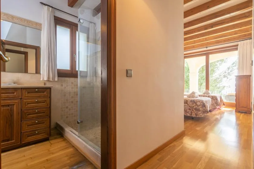 A bathroom with a glass shower and wooden vanity opens onto a bright bedroom.