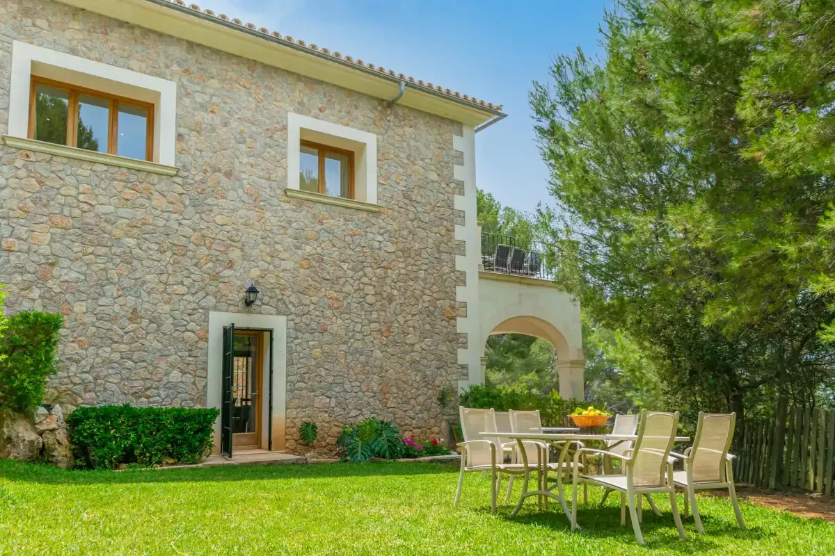 A stone house stands beside a lawn with outdoor seating and trees.
