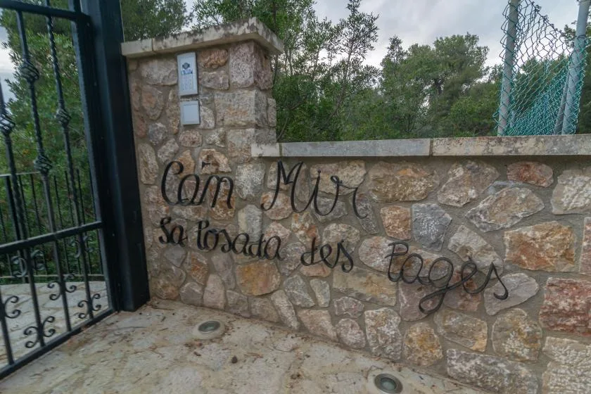 A stone entrance wall with a metal gate and lettering overlooks a wooded area.