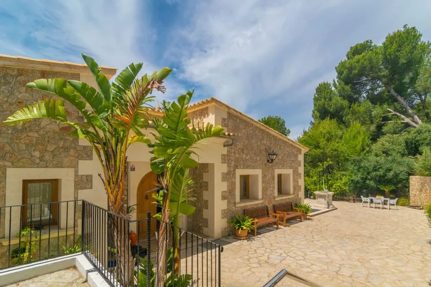 A stone house opens onto a paved patio with outdoor seating and leafy plants.