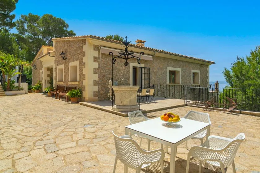 A stone-paved outdoor terrace with a white dining table and seating beside a house.