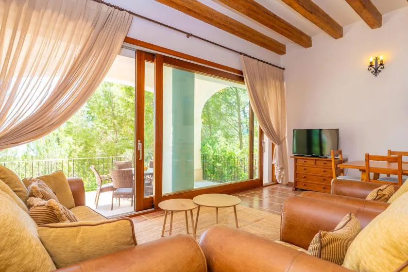A living room with seating, wooden ceiling beams, and glass doors opening to greenery.