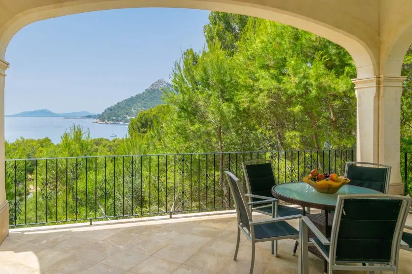 A covered outdoor terrace with a table and seating overlooks trees and the water.