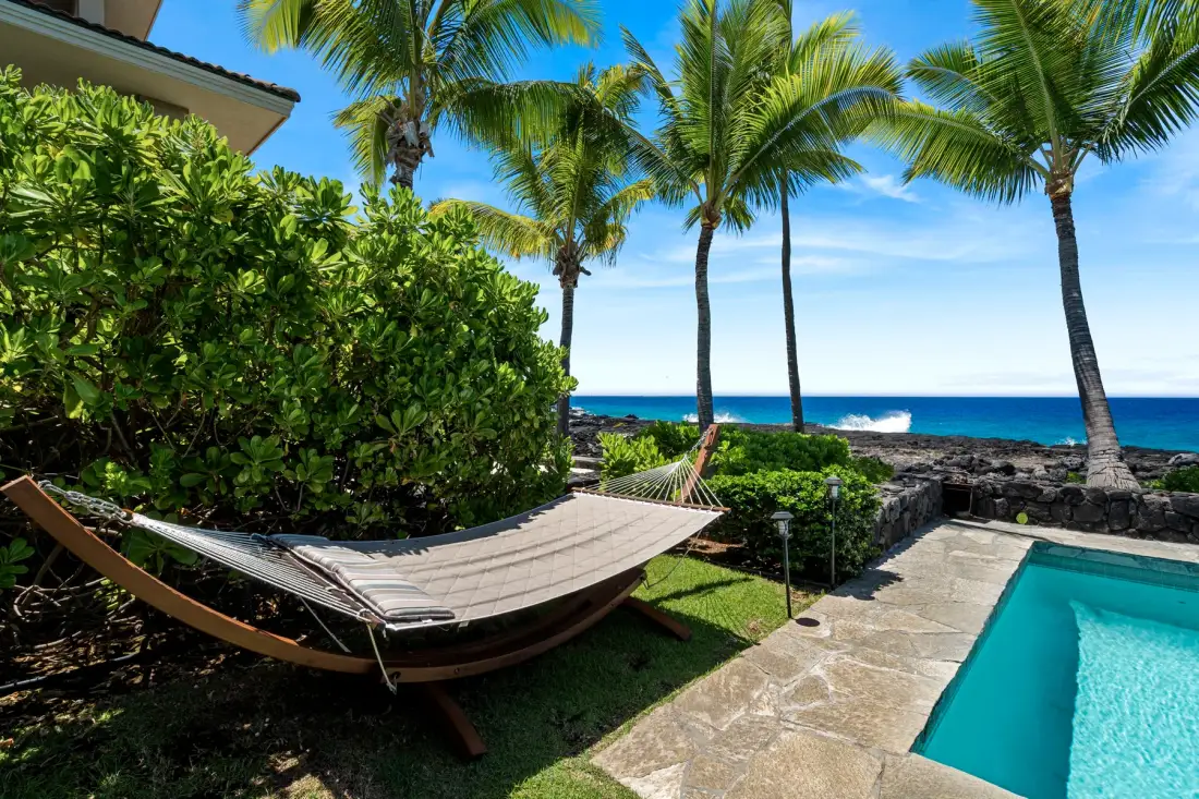 Una zona junto a la piscina con una hamaca, palmeras y vistas al mar.