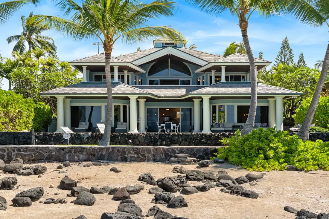 Una gran casa tropical da a una orilla arenosa salpicada de rocas oscuras.