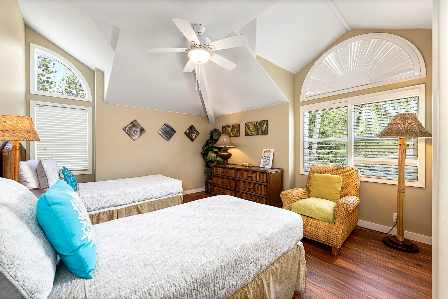 Un dormitorio luminoso con dos camas, un sillón de mimbre y ventanas grandes.