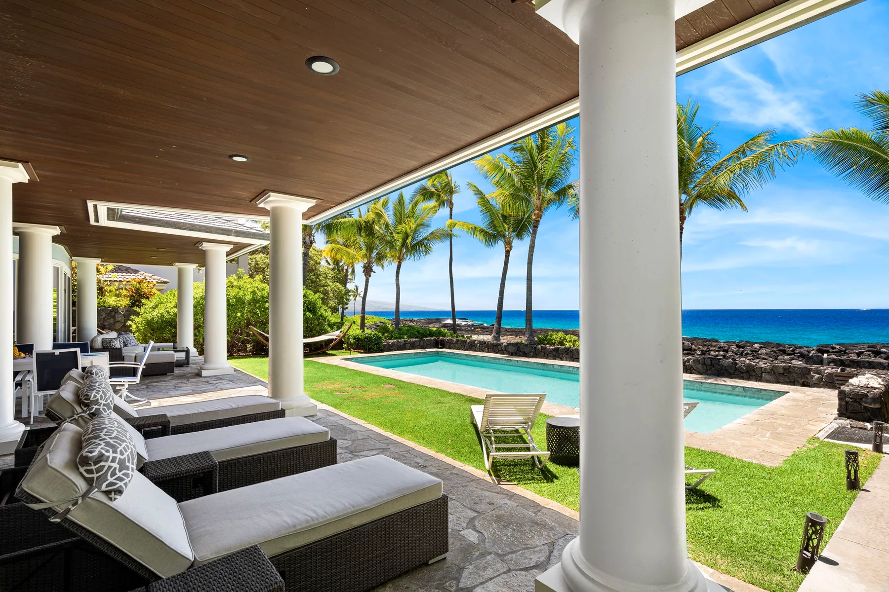 Un patio cubierto con asientos al aire libre da a una piscina, palmeras y el mar.