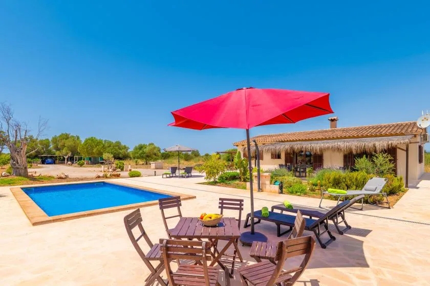 A sunny outdoor patio with a swimming pool, seating, and a red umbrella.