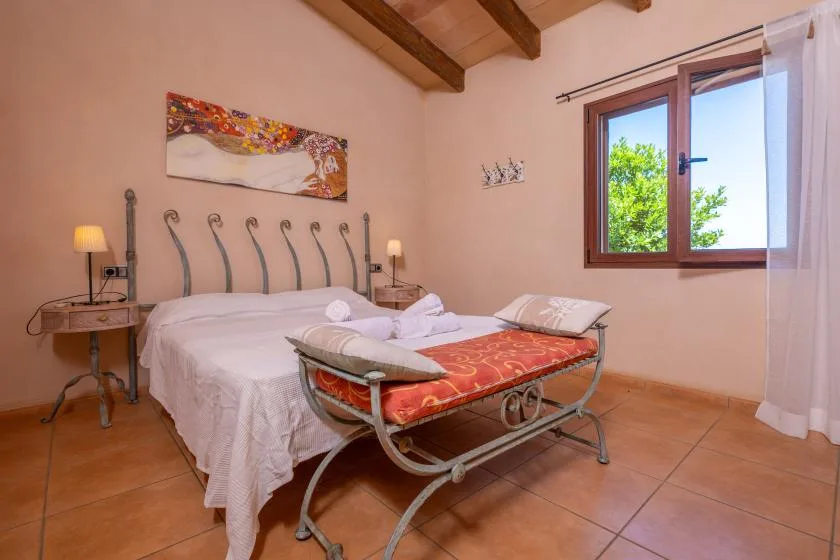 A bedroom with a bed, a red upholstered bench, and a window overlooking greenery.