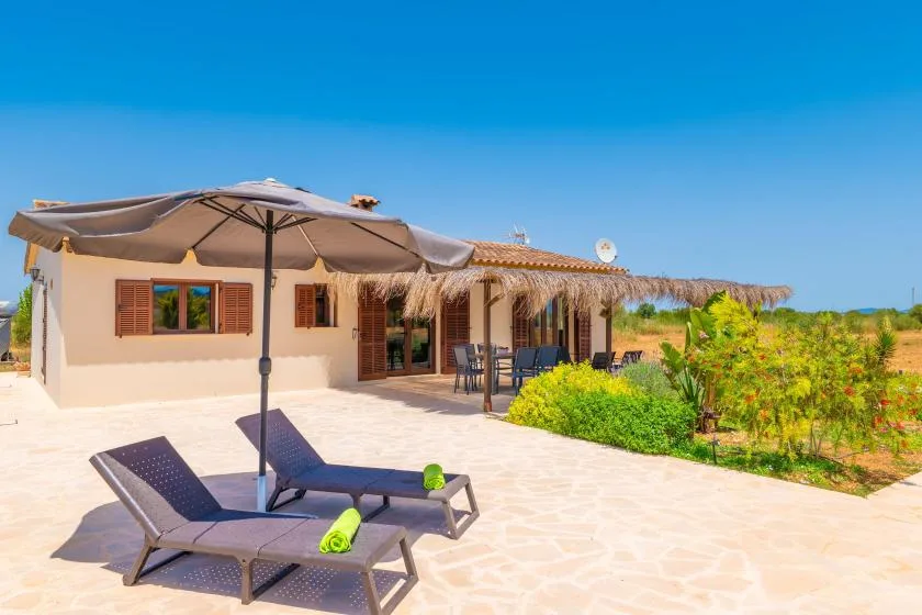A sunny outdoor patio with lounge seating, a large umbrella, and a house behind it.