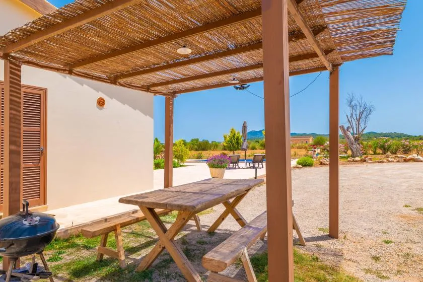 A shaded outdoor dining area with a wooden picnic table beside a white building.