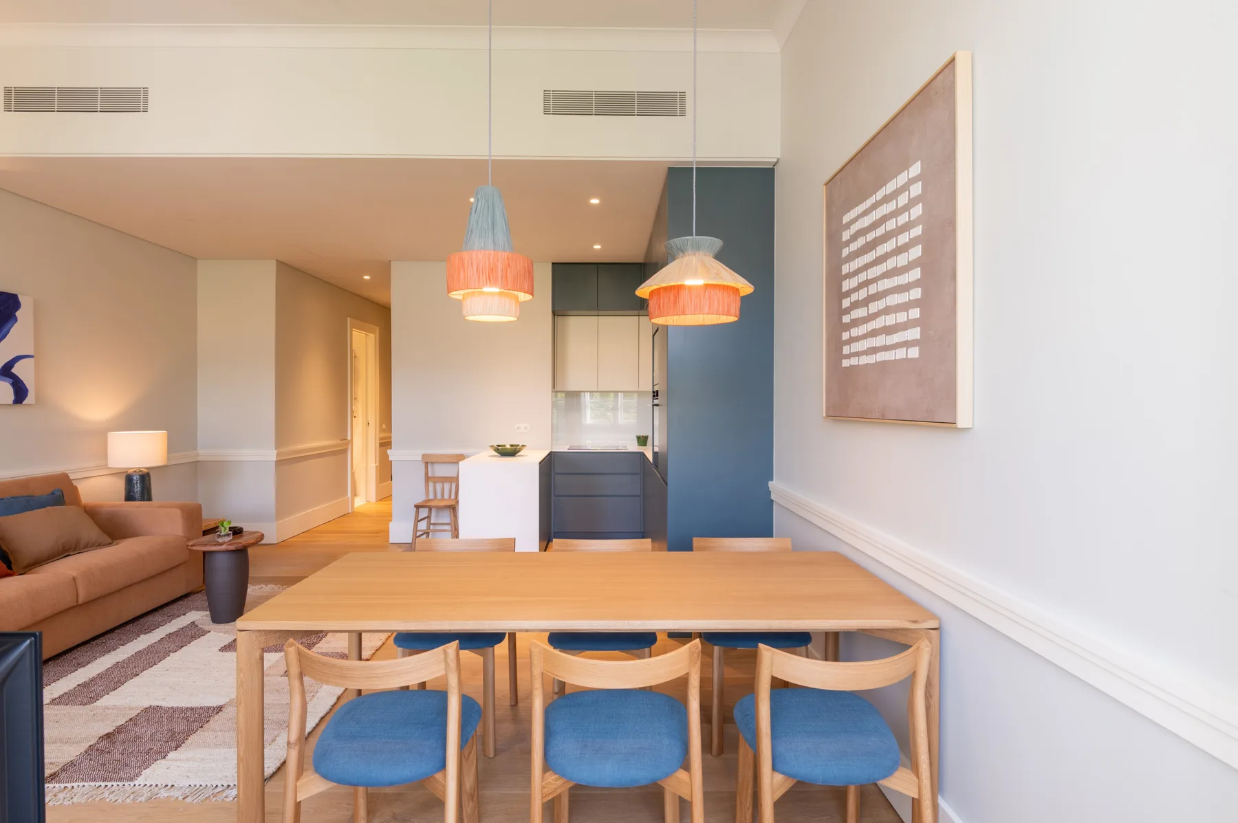 A dining area with a wooden table and blue-cushioned seating opens onto a living area and kitchen.