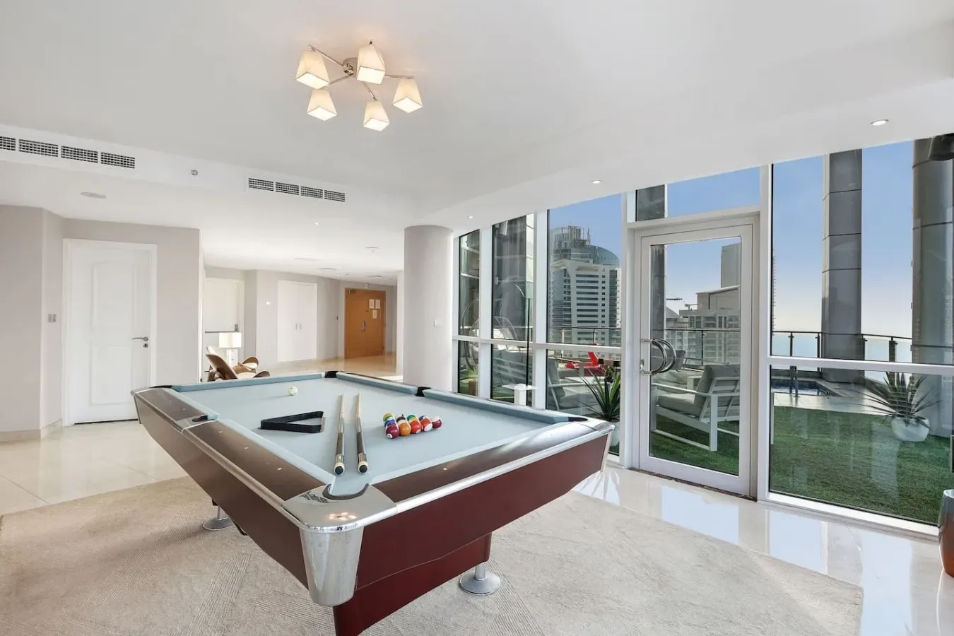 Una sala luminosa tiene una mesa de billar junto a grandes ventanas con vista a edificios cercanos.